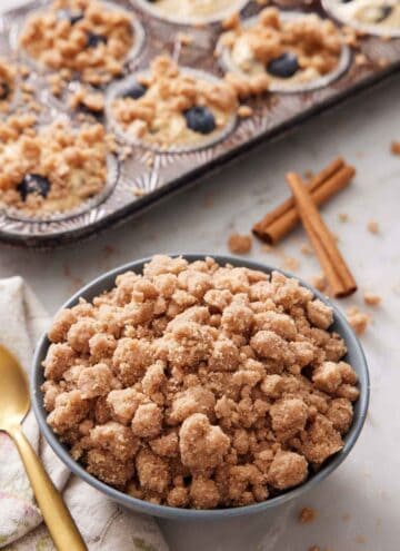 A bowl of streusel topping with cinnamon sticks and muffins topped with streusel in the background.