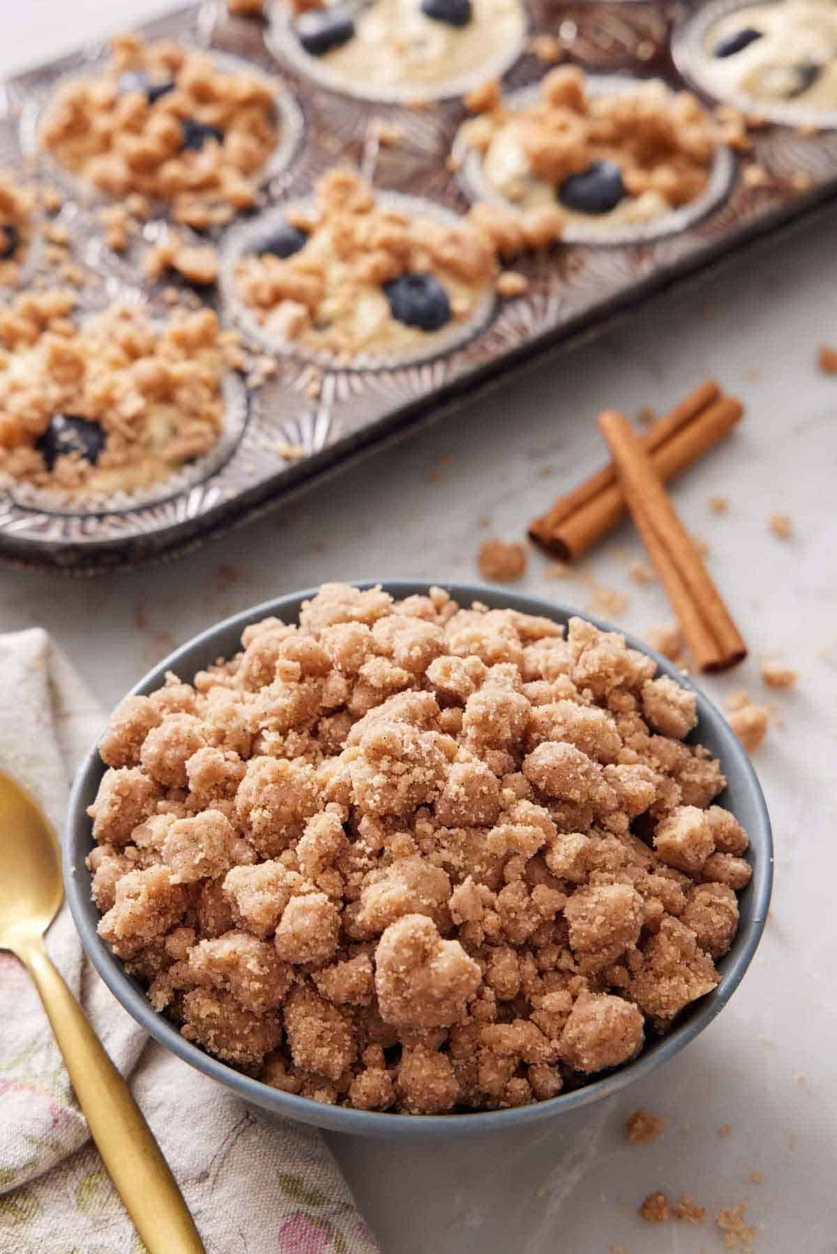 A bowl of streusel topping with cinnamon sticks and muffins topped with streusel in the background.