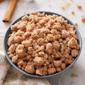 A bowl of streusel topping with some more scattered on the counter along with two cinnamon sticks and a napkin.