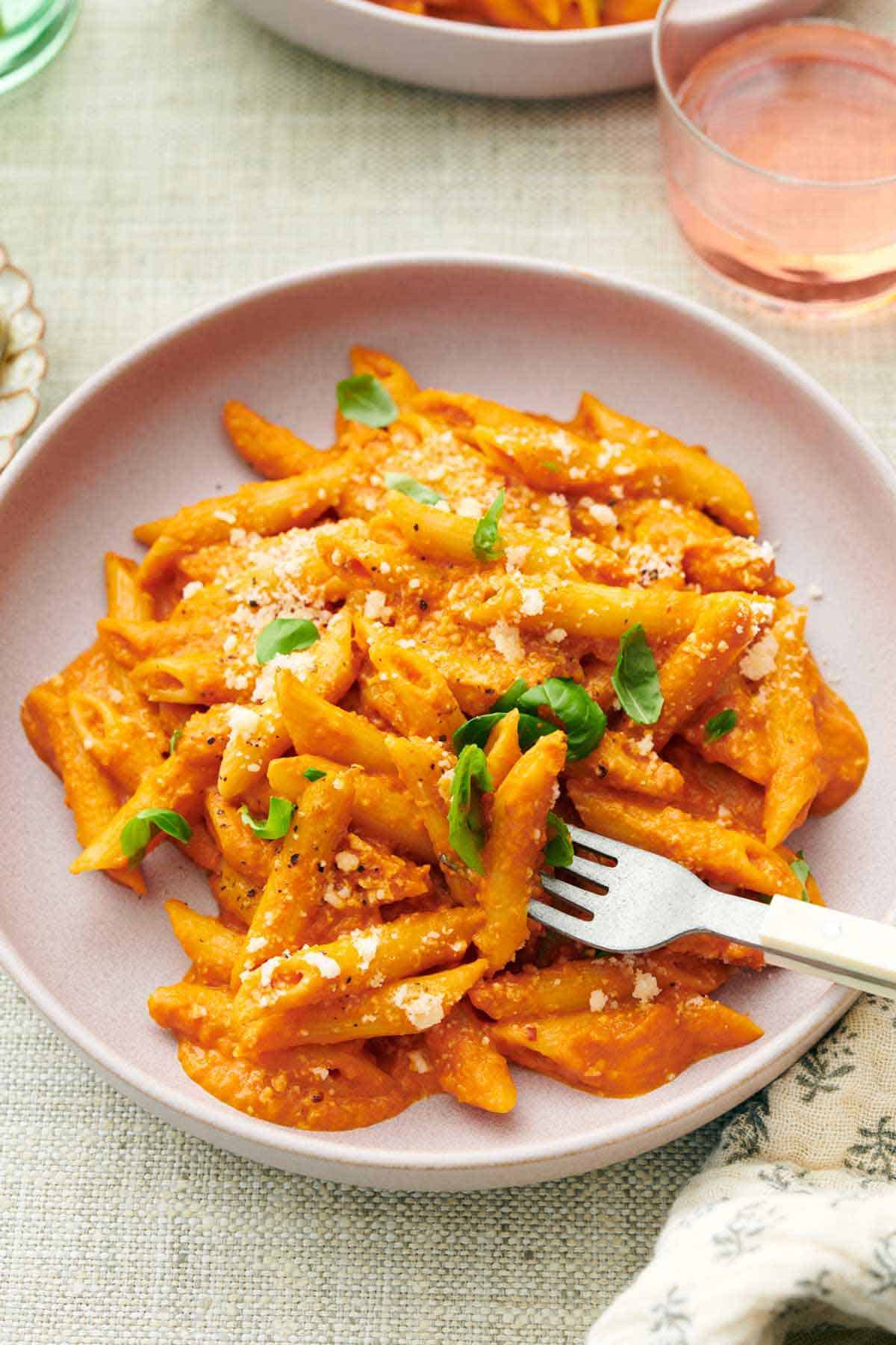 A plate of pasta coated in vodka sauce topped with parmesan and basil with a fork piercing some pasta.