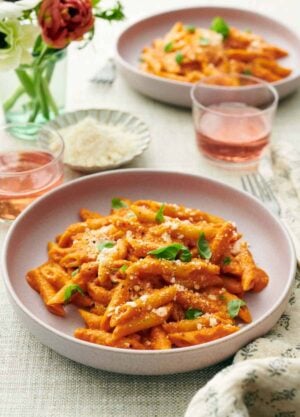 Two plates of pasta coated in vodka sauce topped with cheese and basil. Two drinks, vase of flowers, and a bowl of more parmesan in between the two plates.