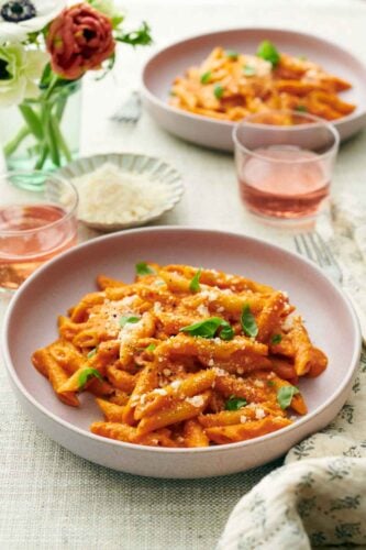 Two plates of pasta coated in vodka sauce topped with cheese and basil. Two drinks, vase of flowers, and a bowl of more parmesan in between the two plates.