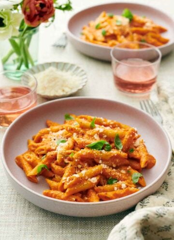 Two plates of pasta coated in vodka sauce topped with cheese and basil. Two drinks, vase of flowers, and a bowl of more parmesan in between the two plates.