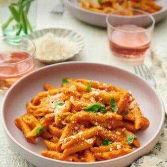 Pinterest graphic of plates of pasta coated in vodka sauce topped with cheese and basil. Two drinks, vase of flowers, and a bowl of more parmesan in between the two plates.