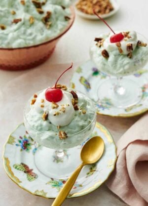 Two dessert bowls on plates with Watergate salad topped with whipped cream, pecans, and a maraschino cherry. A bowl of more Watergate salad in the background.