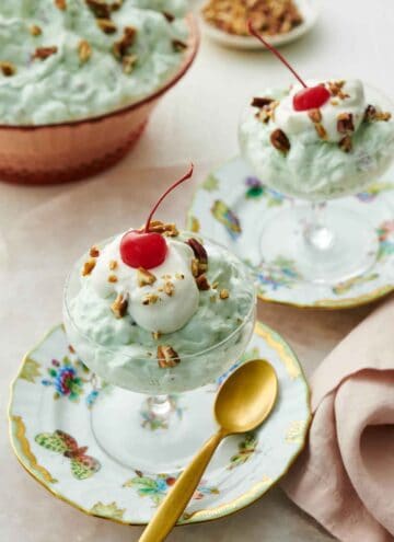 Two dessert bowls on plates with Watergate salad topped with whipped cream, pecans, and a maraschino cherry. A bowl of more Watergate salad in the background.