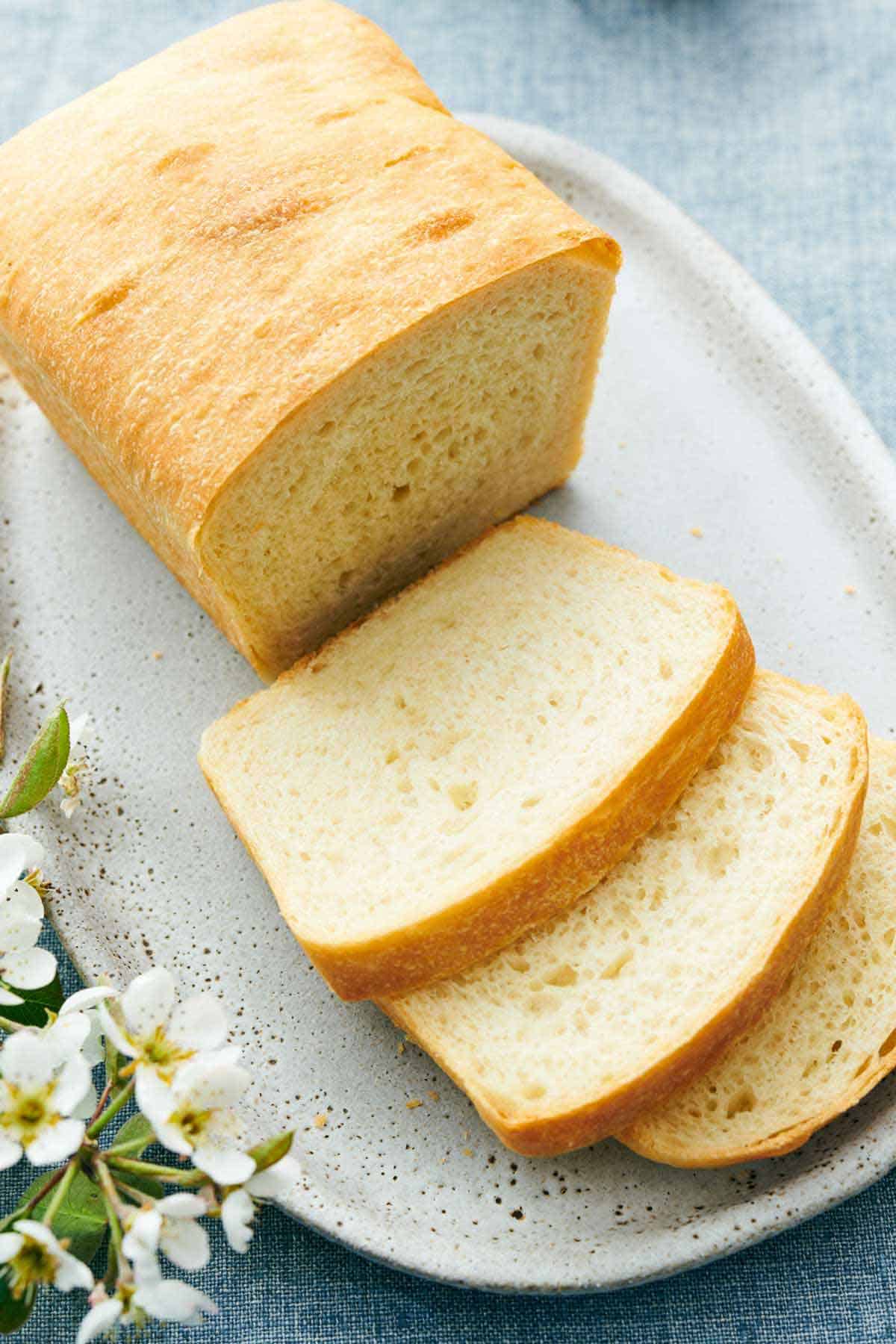 A slightly overhead view of a loaf of white bread with three slices cut in front. Flowers on the side.