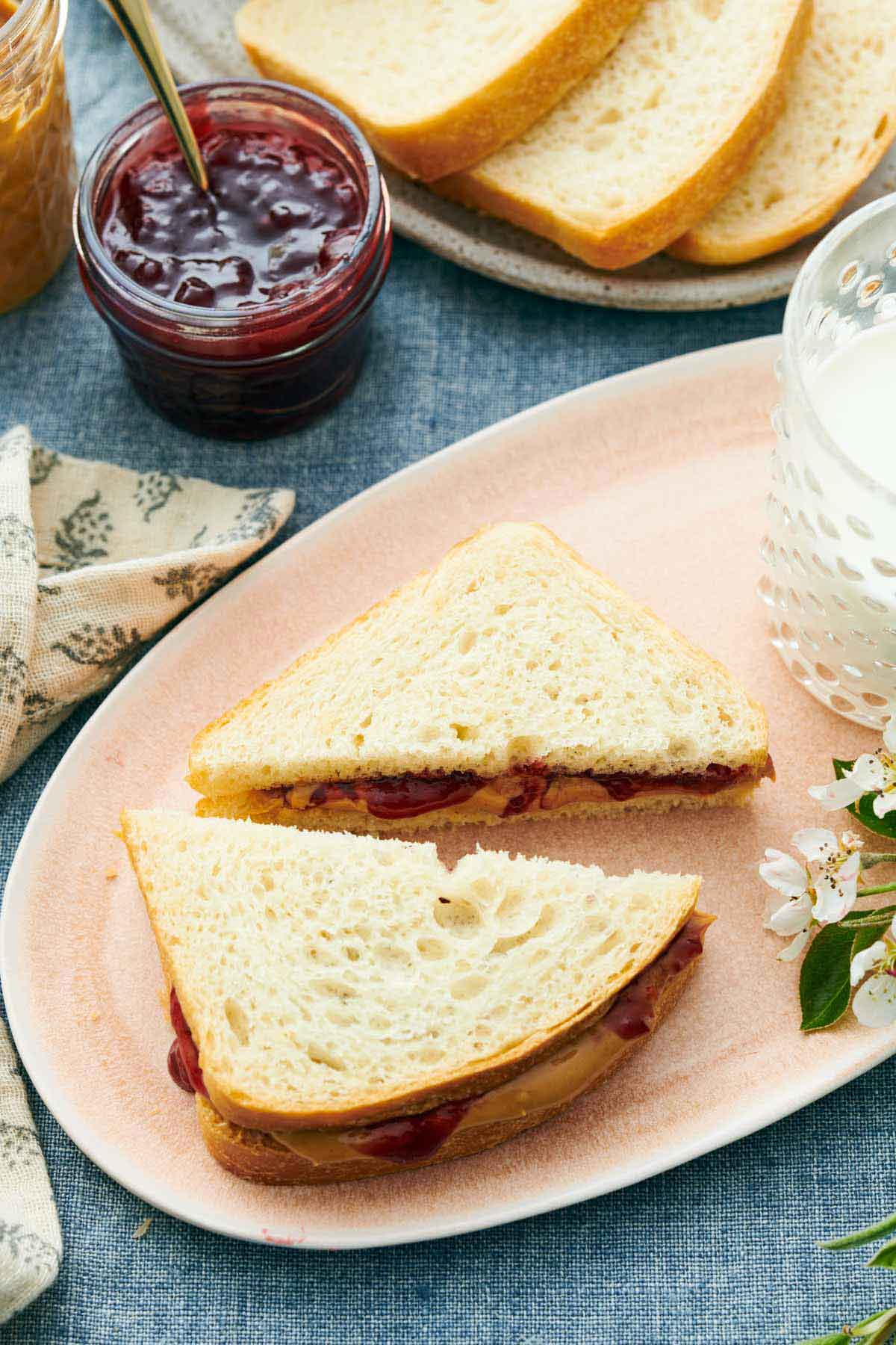 A platter with a peanut butter and jam sandwich made with white bread, cut into two triangles. Jam, bread, and peanut butter on the side.