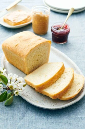 A platter with a loaf of white bread with three slices cut in front. Peanut butter and jam in the background along with a slice of bread with peanut butter.