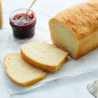 A loaf of white bread on a sheet of parchment paper with two slices cut in front. A jar of jam and peanut butter in the back.