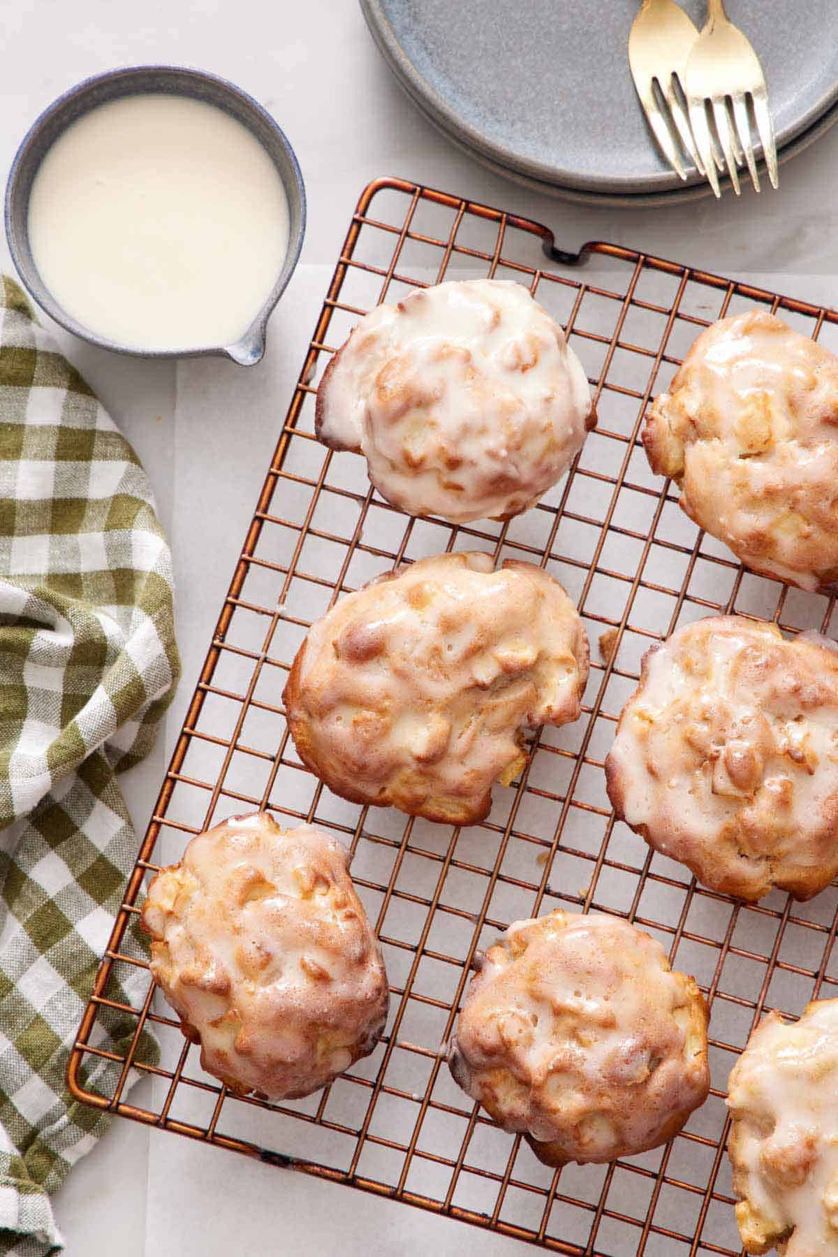 Overhead view of air fryer apple fritters on a cooling rack. A bowl of glaze on the side along with a linen napkin, plates, and forks.