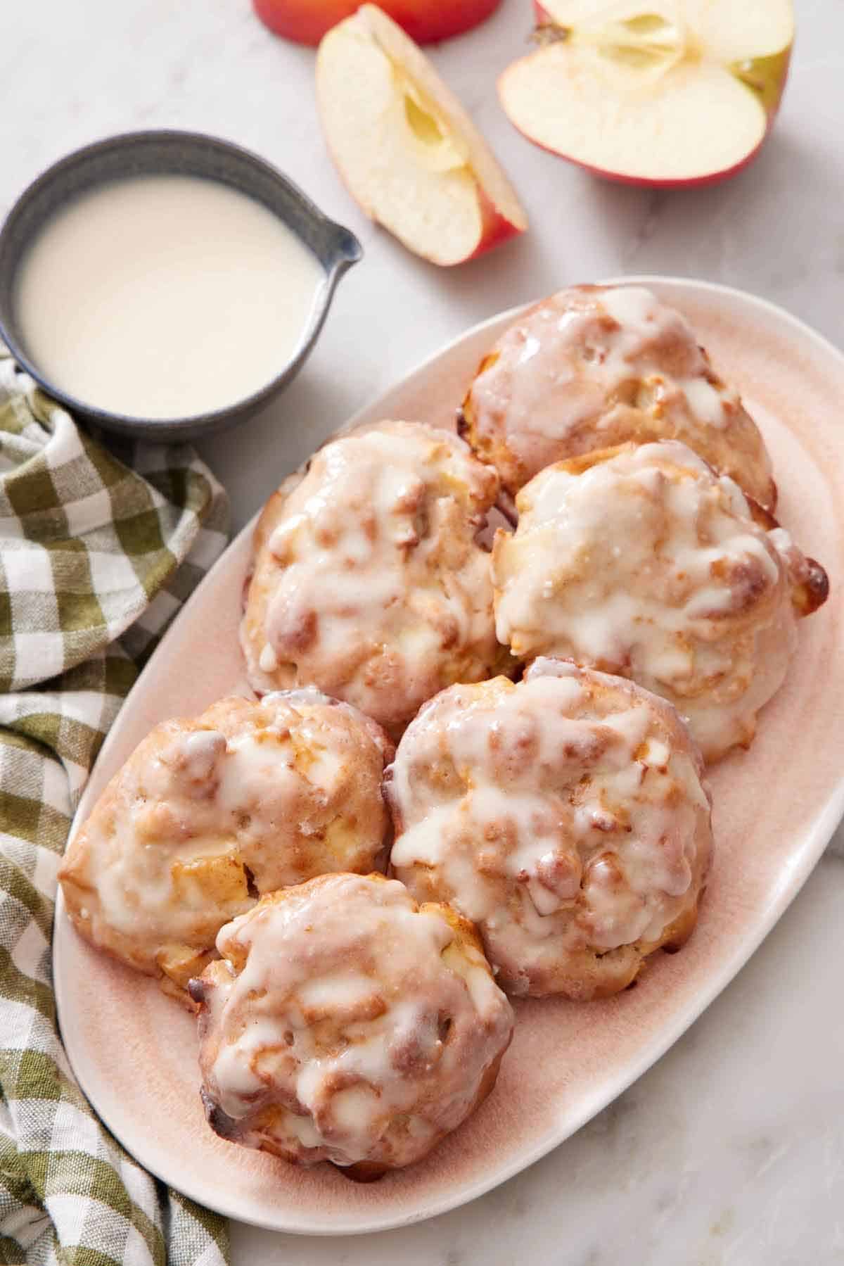 Overhead view of a platter with six air fryer apple fritters. A bowl of glaze and cut apples on the side.