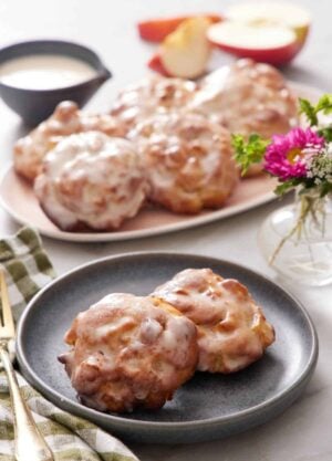 A plate with two pieces of air fryer apple fritters. More fitters on a platter in the background. Cut apples, flowers, and a bowl of glaze in the background.