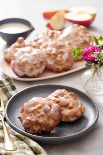 A plate with two pieces of air fryer apple fritters. More fitters on a platter in the background. Cut apples, flowers, and a bowl of glaze in the background.