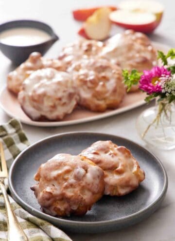 A plate with two pieces of air fryer apple fritters. More fitters on a platter in the background. Cut apples, flowers, and a bowl of glaze in the background.