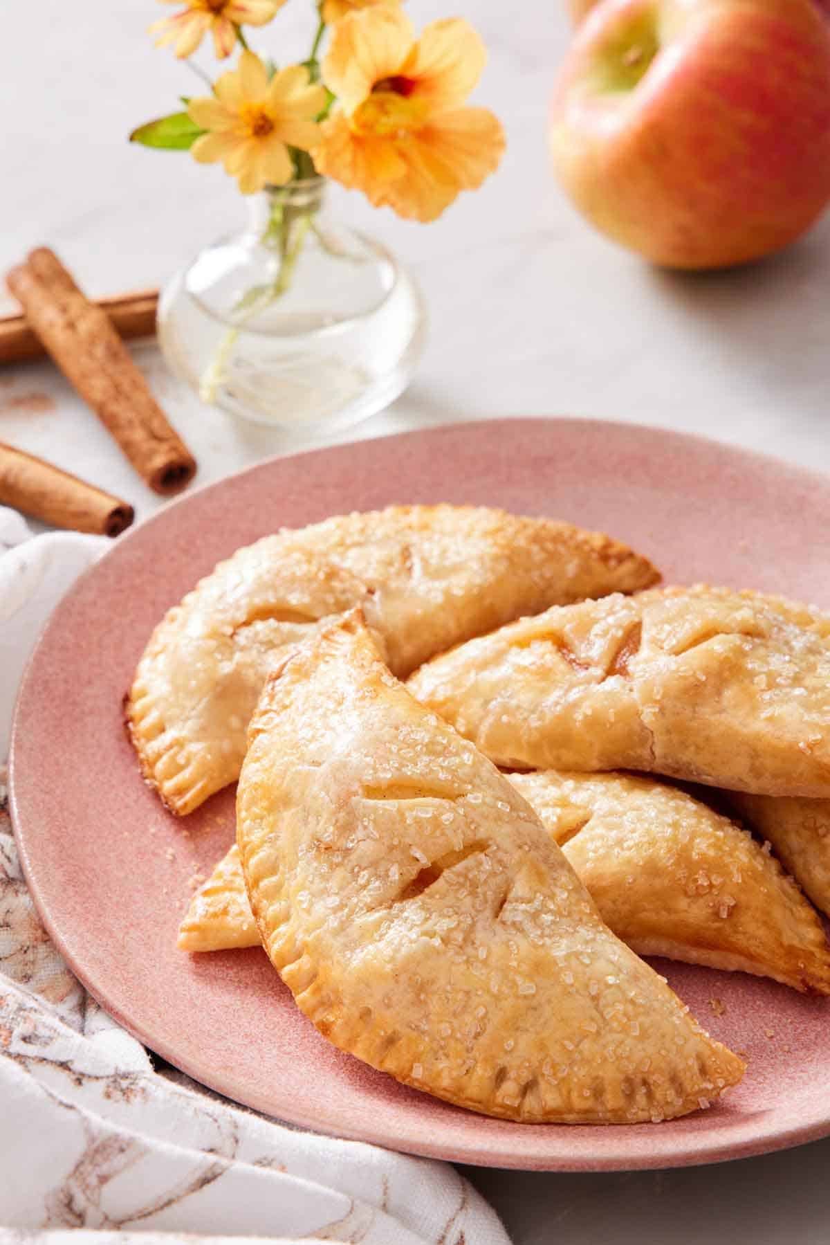 A pink plate with air fryer hand pies. Flowers, apples, and cinnamon sticks in the background.