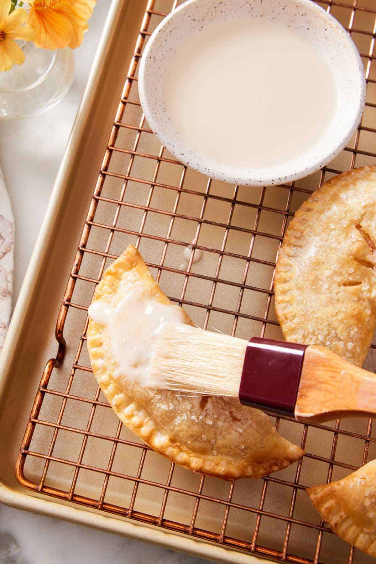 Glaze brushed over an air fryer hand pie on a cooling rack. A bowl of glaze and more hand pies off to the side.