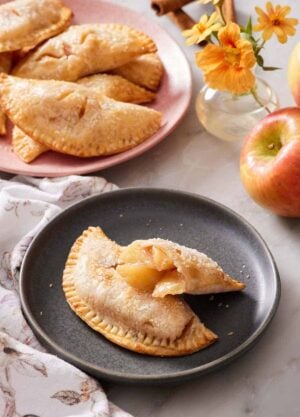 A plate with an air fryer hand pie with a second half leaning on top, showing the interior. More hand pies in the background with flowers, apples, and cinnamon sticks.