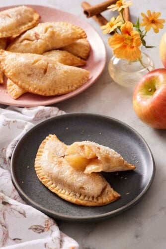 A plate with an air fryer hand pie with a second half leaning on top, showing the interior. More hand pies in the background with flowers, apples, and cinnamon sticks.