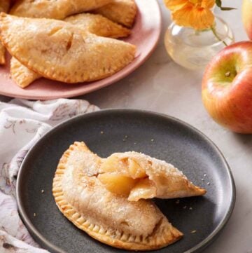 A plate with an air fryer hand pie with a second half leaning on top, showing the interior. More hand pies in the background with flowers, apples, and cinnamon sticks.