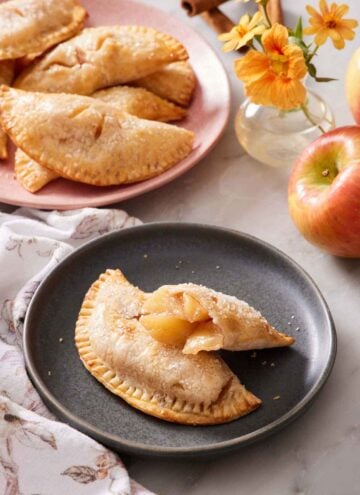 A plate with an air fryer hand pie with a second half leaning on top, showing the interior. More hand pies in the background with flowers, apples, and cinnamon sticks.