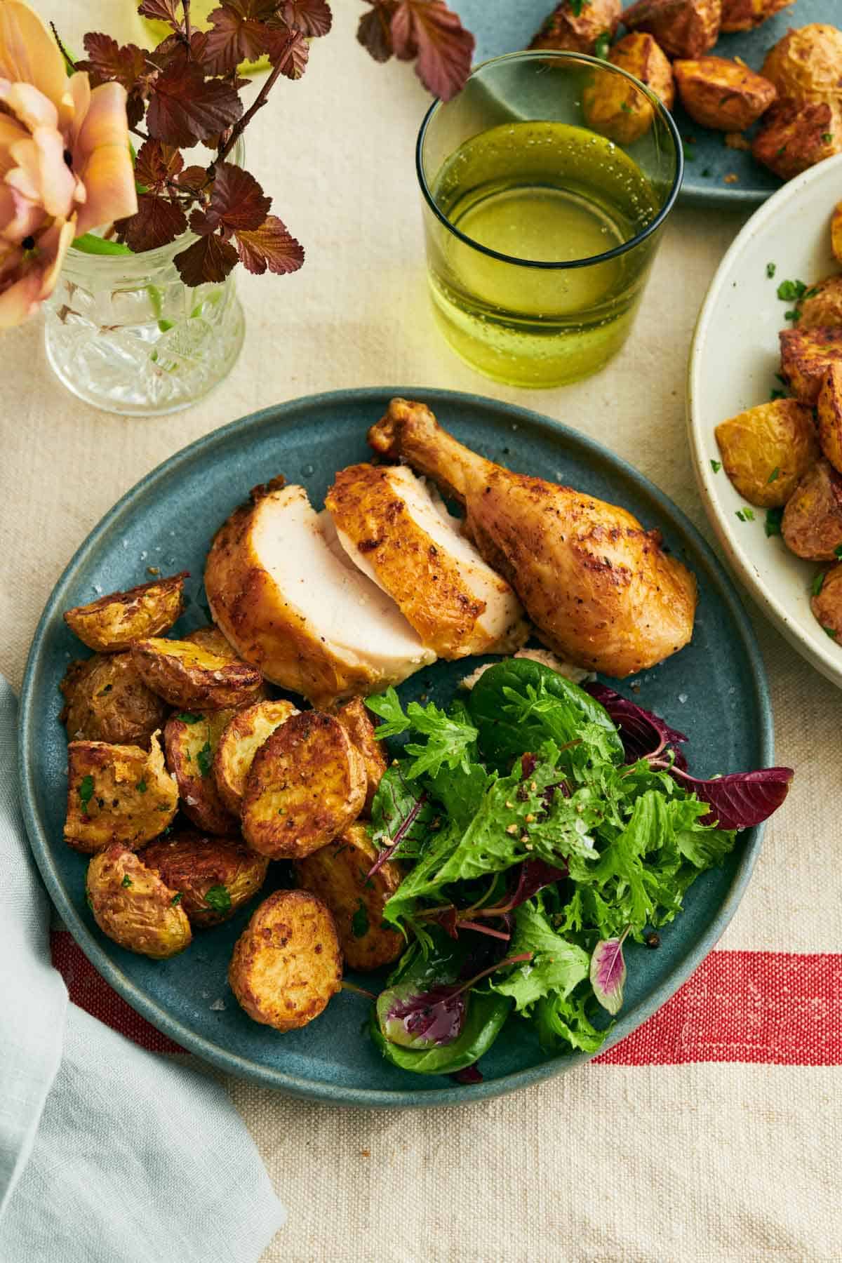 A plate of air fryer potatoes, salad, and chicken. A drink and a vase of flowers off to the side with more potatoes.