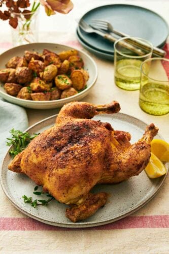 A platter with air fryer whole chicken with some herbs and lemon wedges. Two glasses of water and baby potatoes in the background along with plates and forks.