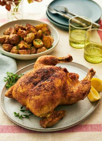 A platter with air fryer whole chicken with some herbs and lemon wedges. Two glasses of water and baby potatoes in the background along with plates and forks.