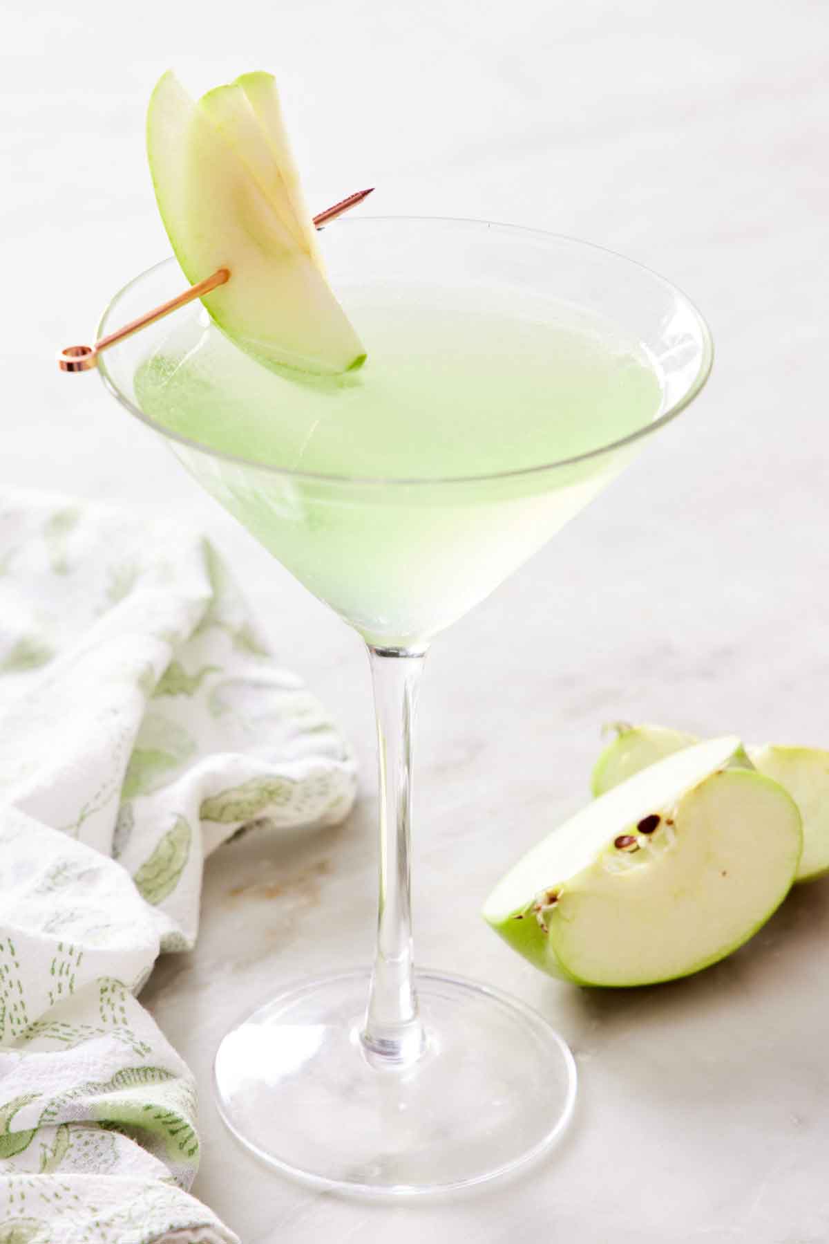 A glass of apple martini with sliced apples on a cocktail pick. Cut apples on the side.