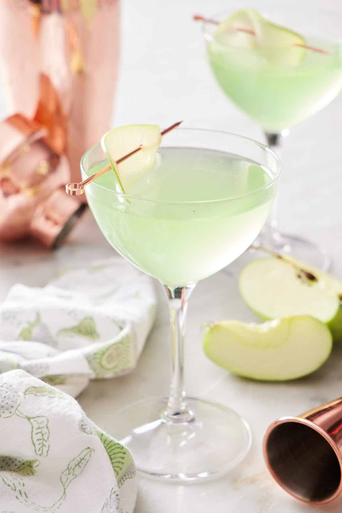 Two glasses of apple martini. A jigger, cut apples, cocktail shaker, and linen napkin on the side.