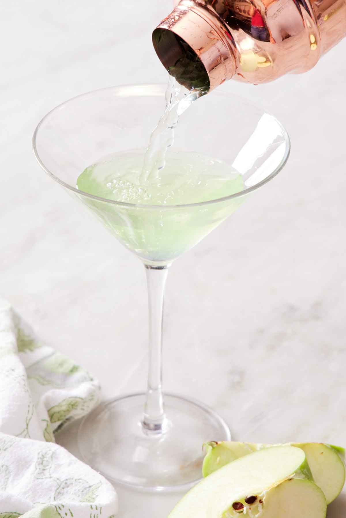 A glass with an apple martini poured in from a cocktail shaker. Cut apples and linen napkin in the front.