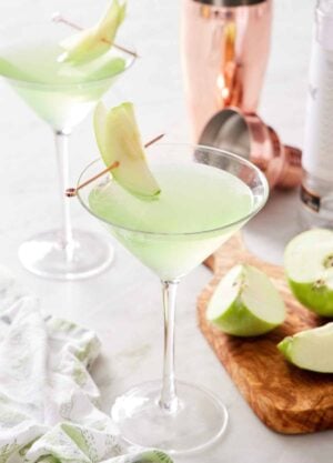 Two glasses of apple martini with sliced apple garnishes. Cut apples and a cocktail shaker on the side.