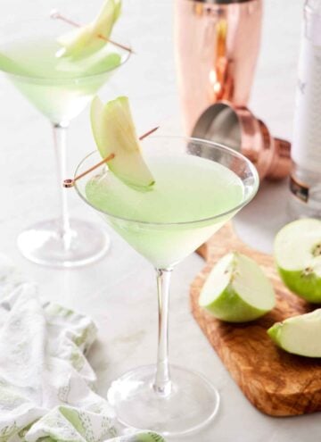 Two glasses of apple martini with sliced apple garnishes. Cut apples and a cocktail shaker on the side.