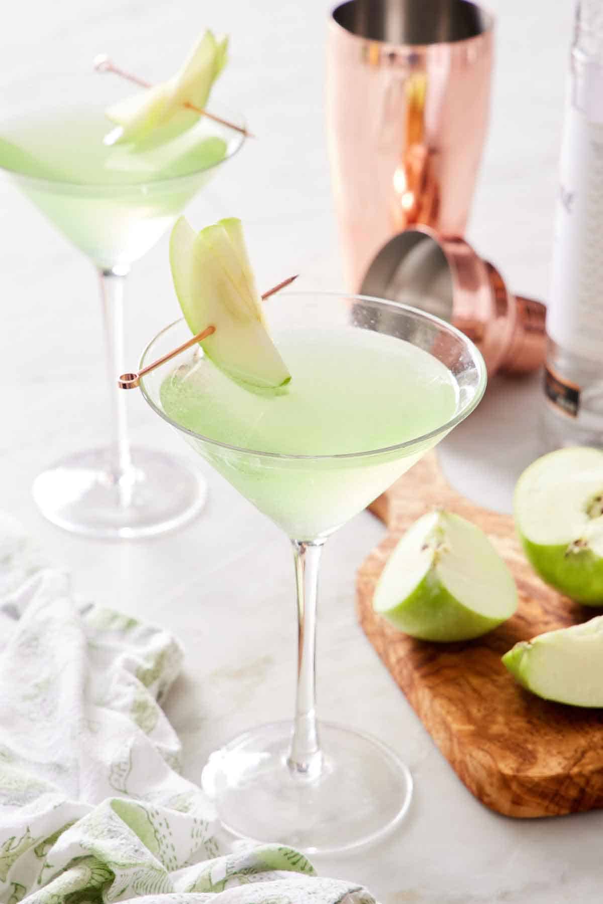 Two glasses of apple martini with sliced apple garnishes. Cut apples and a cocktail shaker on the side.