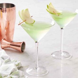 Two glasses of apple martini with sliced apple garnishes. A cocktail shaker and jigger on the side with a napkin.