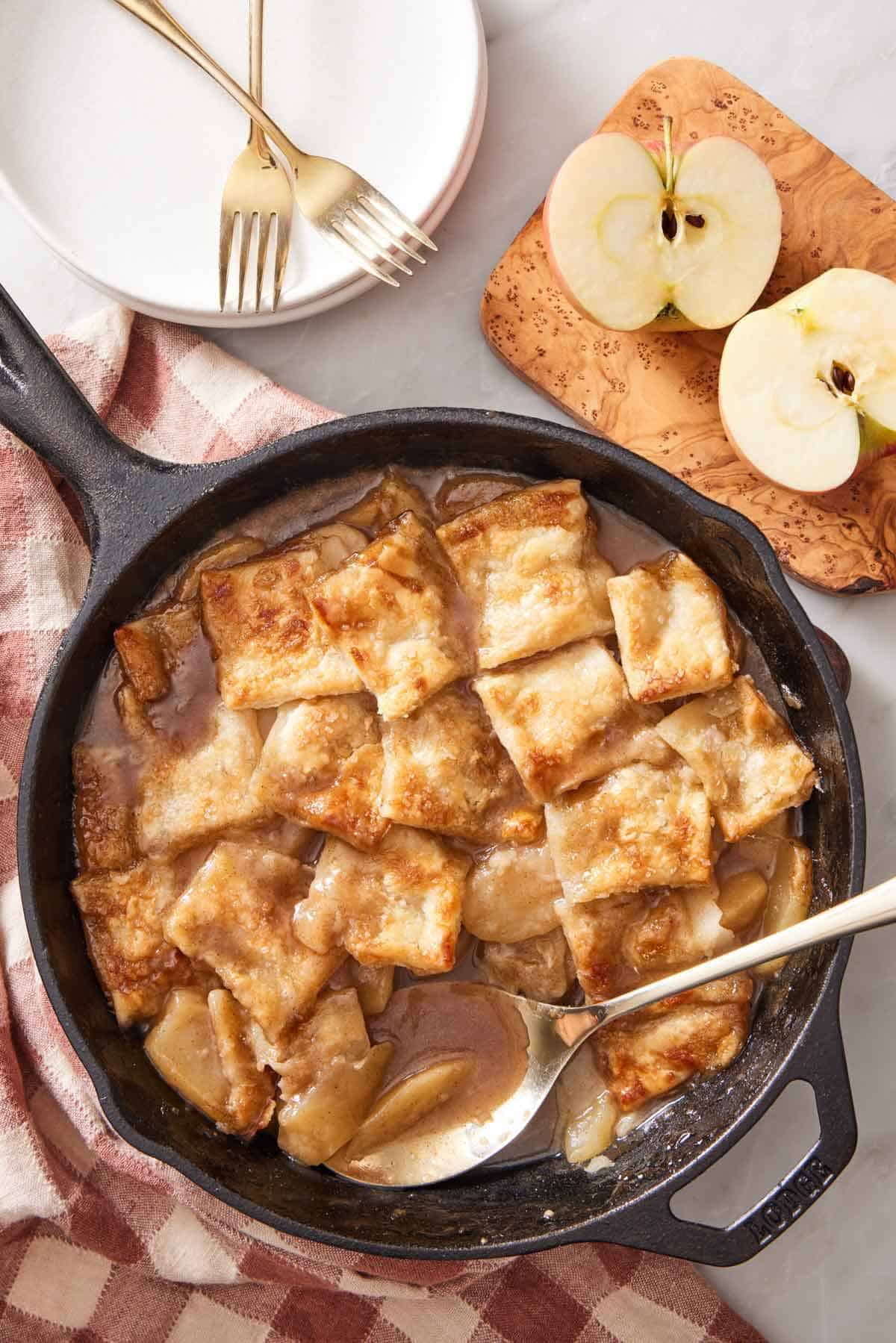 Overhead view of a skillet of apple pandowdy with a spoon tucked in. A cup apple on the side with plates and forks.