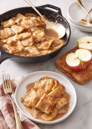 A plate with a serving of apple pandowdy with a skillet in the background containing the rest with a serving spoon. Cut apples on the side.
