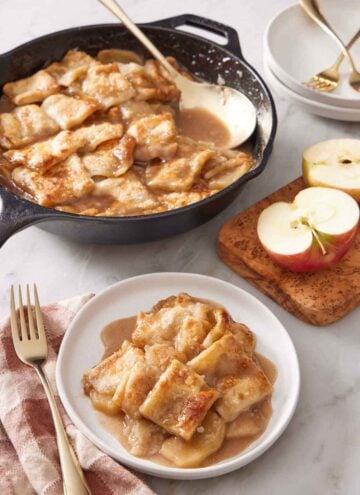 A plate with a serving of apple pandowdy with a skillet in the background containing the rest with a serving spoon. Cut apples on the side.