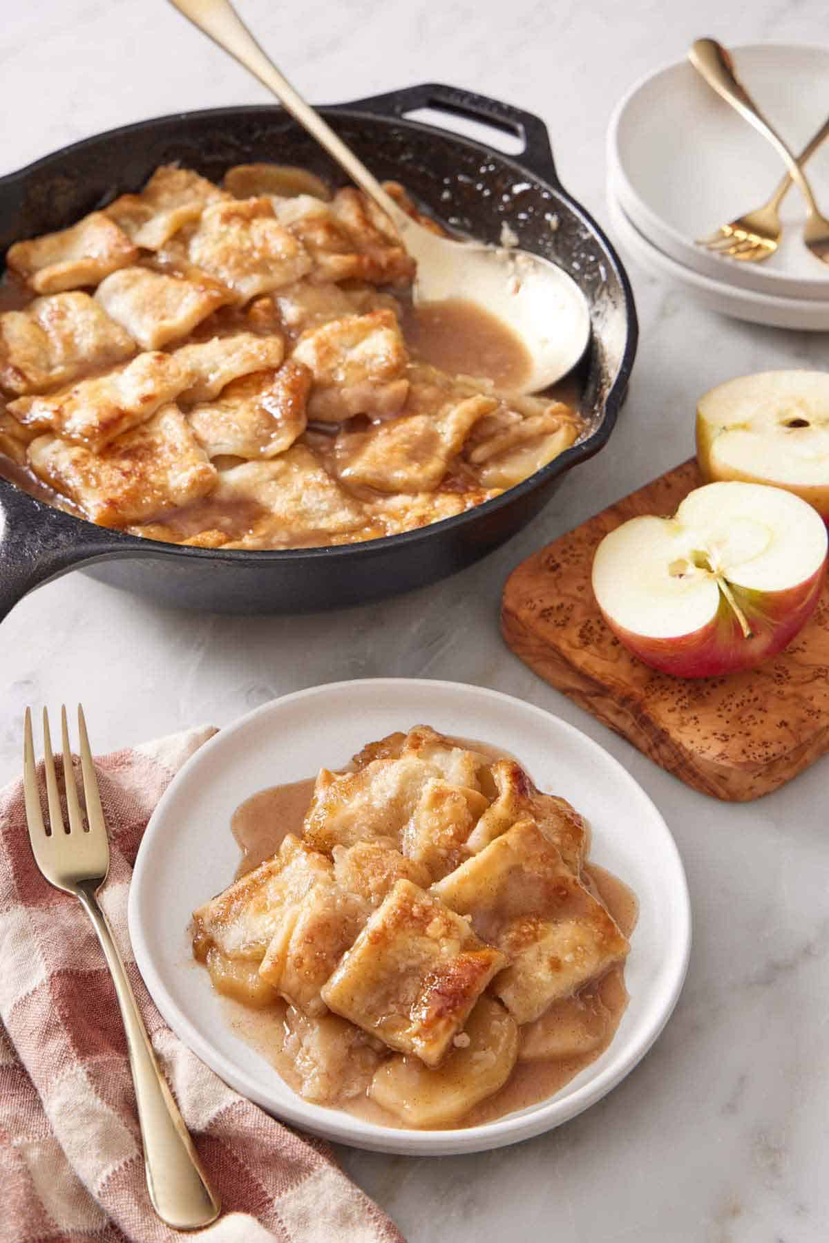 A plate with a serving of apple pandowdy with a skillet in the background containing the rest with a serving spoon. Cut apples on the side.