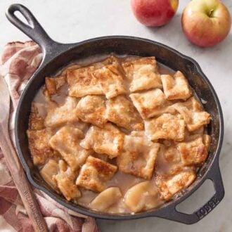 A cast iron skillet of apple pandowdy with a wooden spoon on the side. Two apples off to the side.