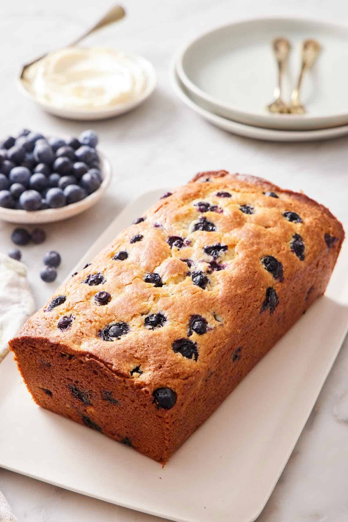 A loaf of blueberry bread on a platter. A bowl of blueberries, plates, and forks in the background.