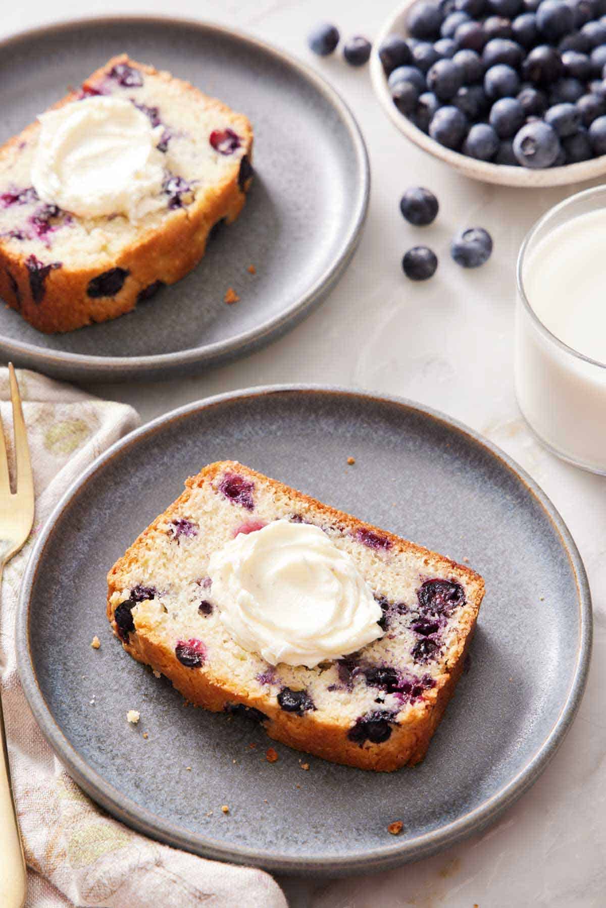 Two plates with sliced blueberry bread with frosting. A glass of milk and bowl of blueberries in the background.