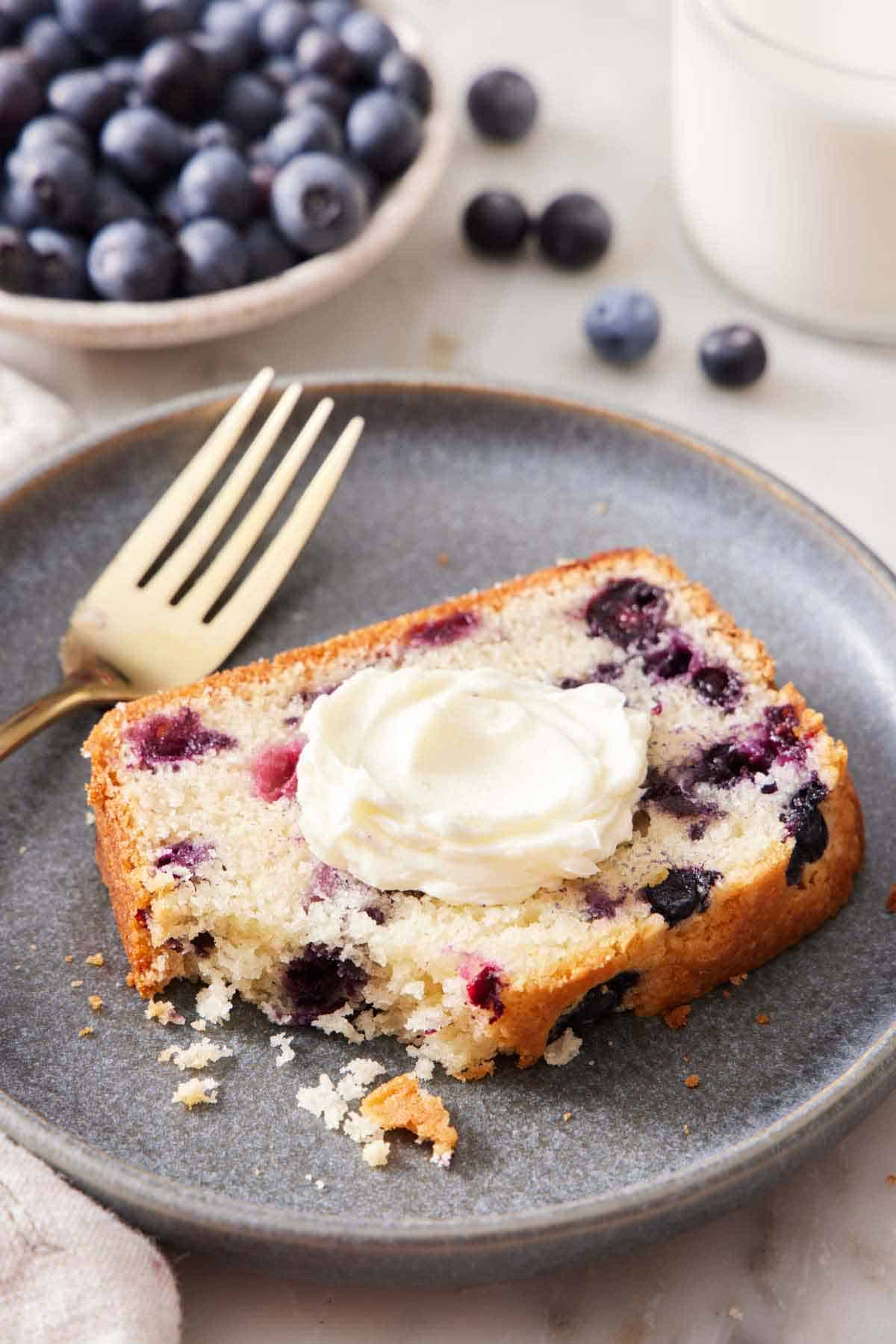 A plate with a slice of blueberry bread with the corner eaten. A fork on the plate. A bowl of blueberries in the back.