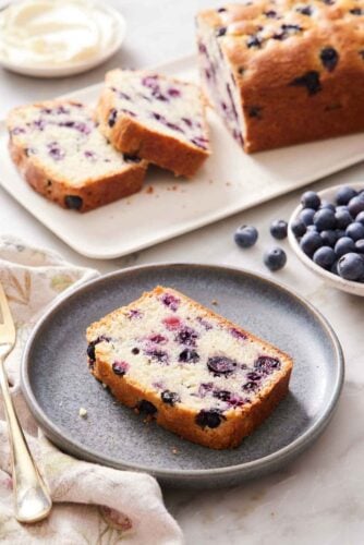 A slice of blueberry bread on a plate with a cut loaf in the background along with a bowl of blueberries.