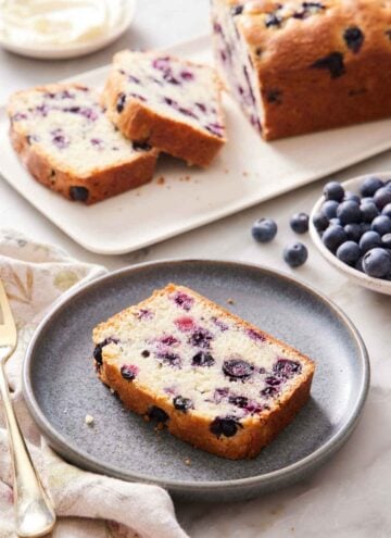 A slice of blueberry bread on a plate with a cut loaf in the background along with a bowl of blueberries.