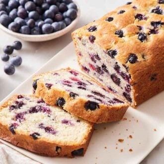 A loaf of blueberry bread with two slices cut in front. A bowl of blueberries in the background.