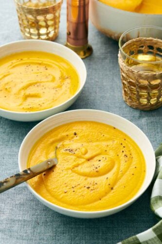 A bowl of carrot soup with a spoon with pepper on top. A second bowl in the back along with drinks.