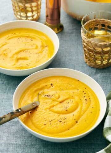 A bowl of carrot soup with a spoon with pepper on top. A second bowl in the back along with drinks.