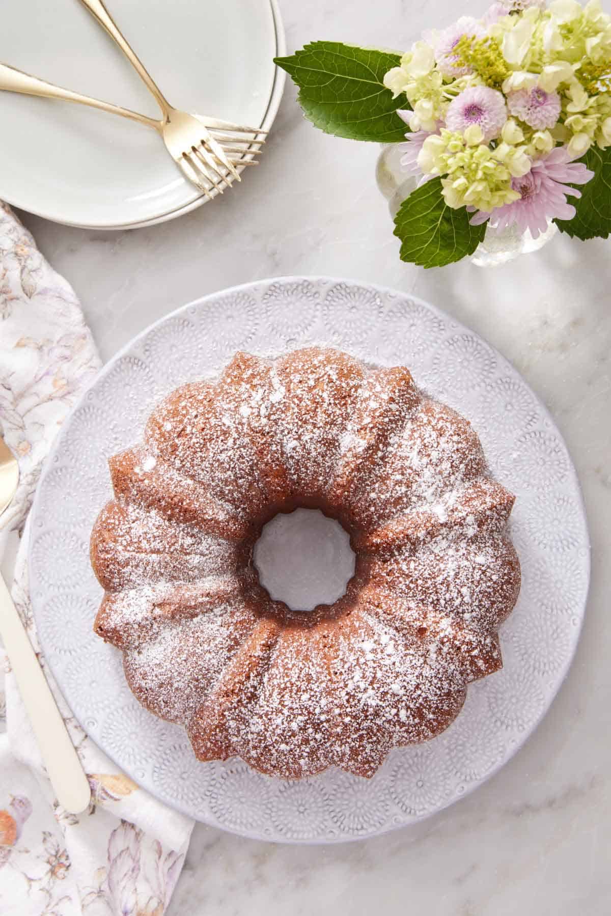 Overhead view of a platter with a cream cheese pound cake dusted with powdered sugar. Flowers, plates, and forks off to the side.