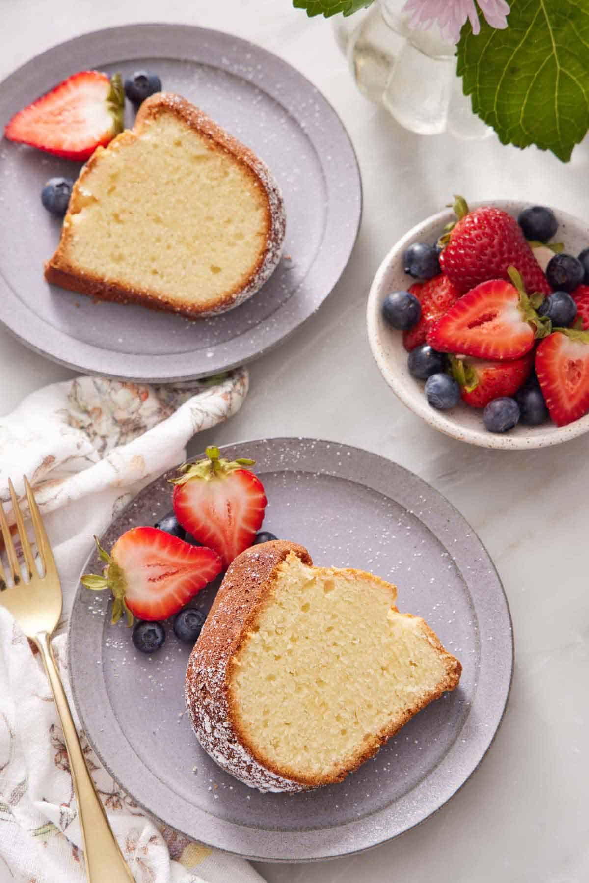 Overhead view of two plates of sliced cream cheese pound cake with berries dusted with powdered sugar. A bowl of berries on the side.